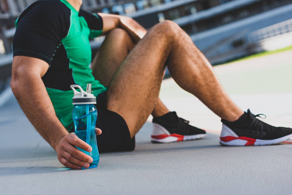 Ausgeschnittene Ansicht eines Sportlers, der auf der Laufbahn sitzt und Flasche mit Wasser in der Hand hält - Foto, Bild