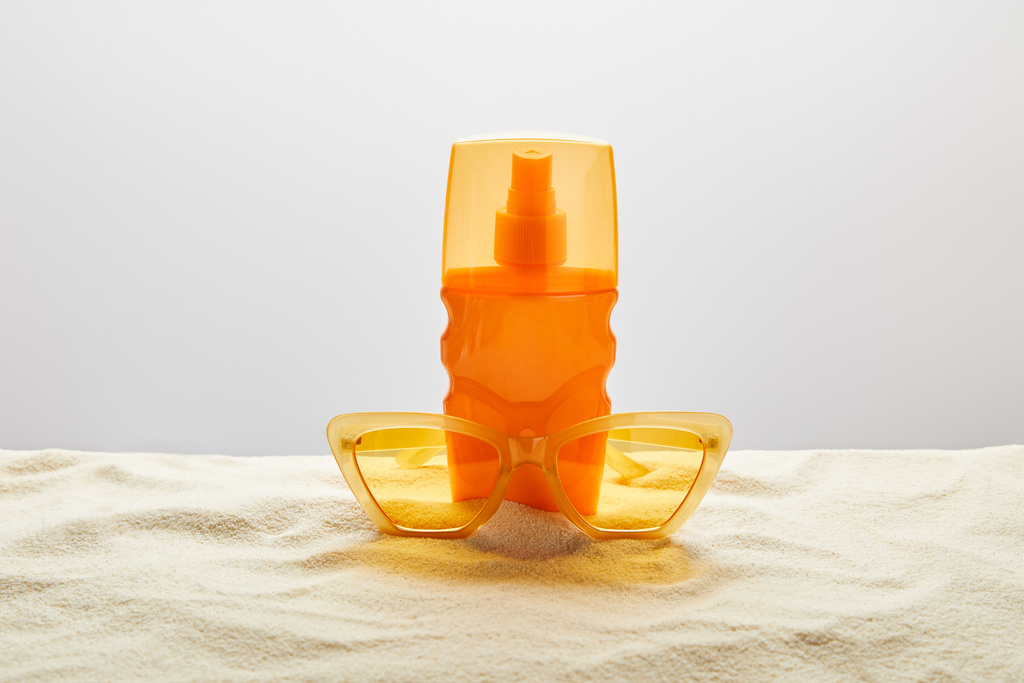 желтые стильные солнцезащитные очки и солнцезащитный крем в оранжевой бутылке на песке с ракушкой на сером фоне
 - Фото, изображение