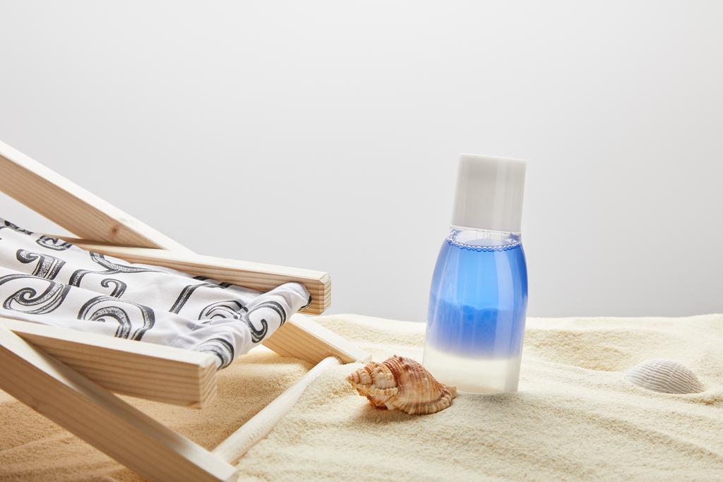 синий лосьон в бутылке в песке возле раковин и шезлонг на сером фоне с копировальным пространством
 - Фото, изображение