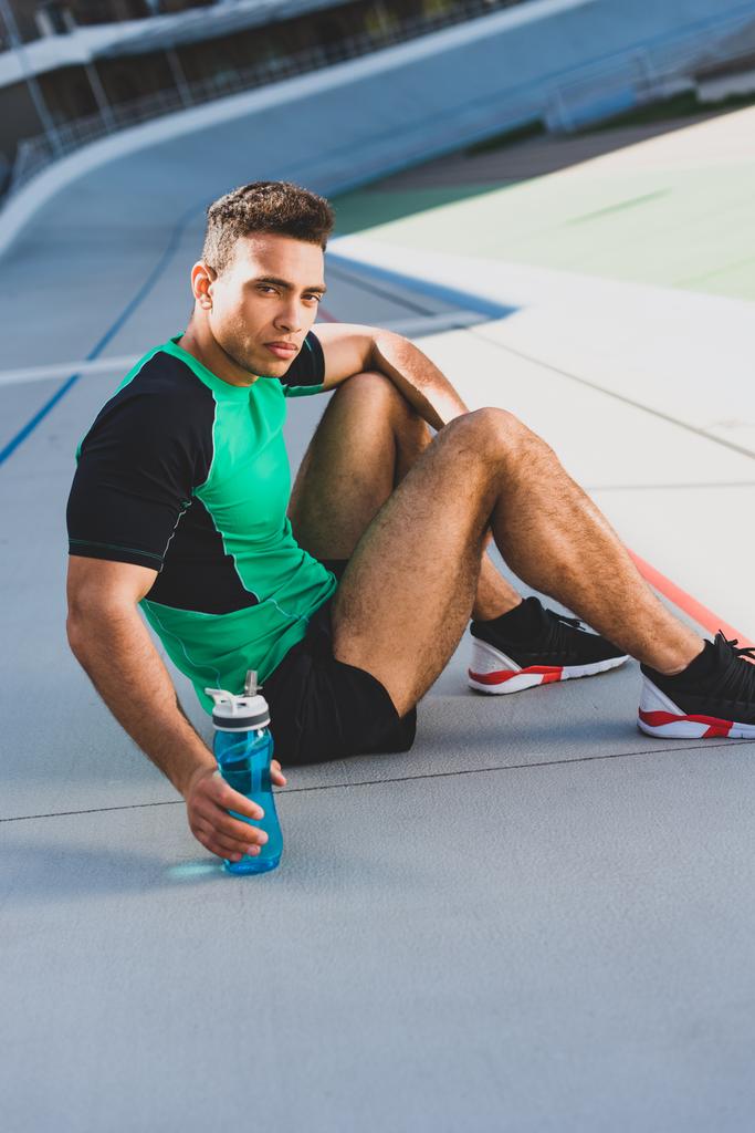 karışık yarış sporcusu koşu pistinde oturuyor, ciddiye kamera bakıyor ve su ile şişe tutarak - Fotoğraf, Görsel