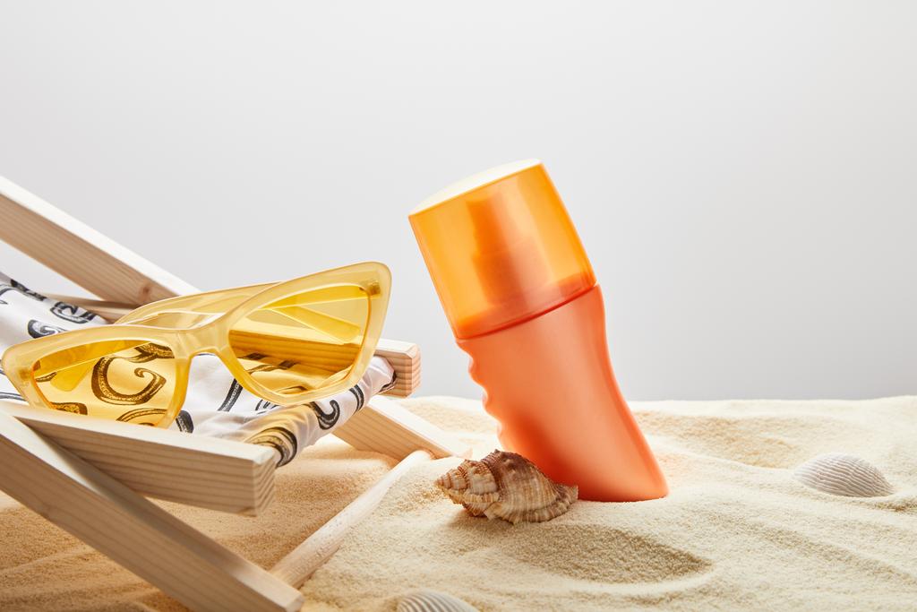 pomarańczowy filtr przeciwsłoneczny w piasku w pobliżu muszli, żółte okulary przeciwsłoneczne i leżak na szarym tle - Zdjęcie, obraz