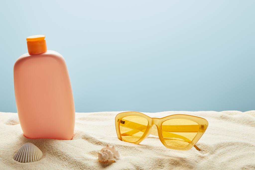 αντηλιακή λοσιόν στην άμμο κοντά σε κοχύλια και κίτρινα γυαλιά ηλίου σε μπλε φόντο - Φωτογραφία, εικόνα