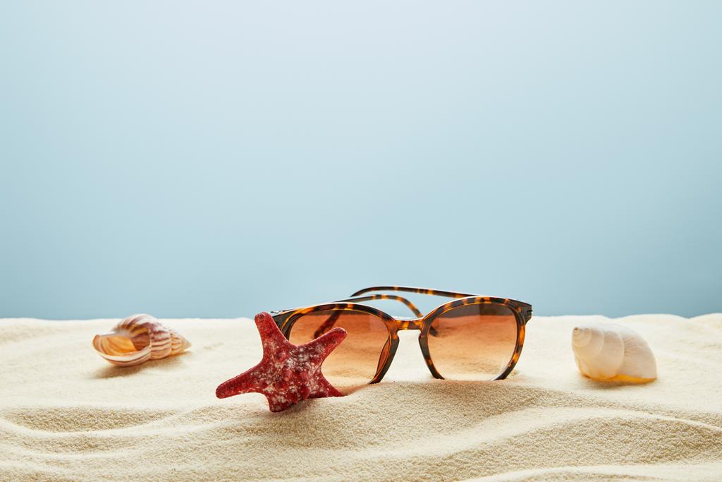 óculos de sol castanhos e elegantes na areia com conchas e estrelas do mar no fundo azul
 - Foto, Imagem