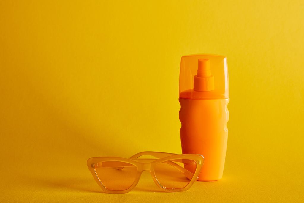 濃い黄色の背景にサングラスの近くにオレンジ色のボトルの日焼け止め - 写真・画像
