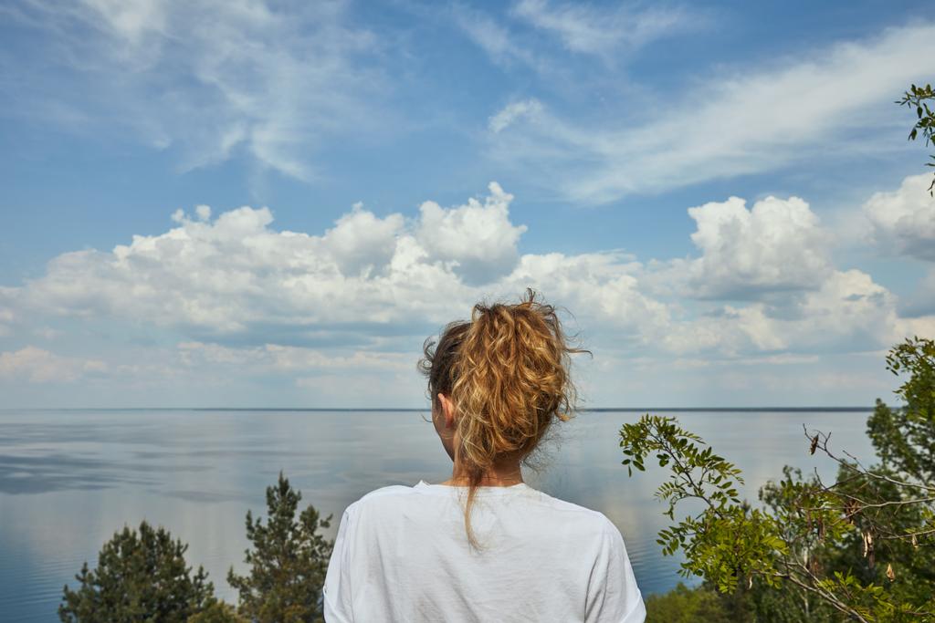 Вид ззаду на молоду жінку, дивлячись на пейзаж з зеленими листочками, річкою і блакитним небом
 - Фото, зображення