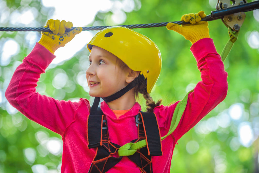 木々の間のロープパーク上の美しい子供の肖像画。夏の幸せな子供。ローピングパーク。ロープパーク - クライミングセンター。幼児の年齢。公園で木に登る子供。すべての子供時代の問題. - 写真・画像