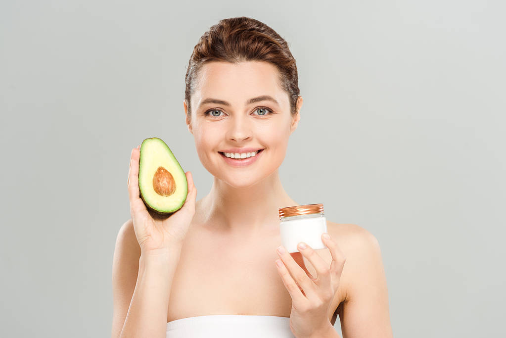 vrolijke vrouw houdt de helft van avocado en container met cosmetische crème geïsoleerd op grijs  - Foto, afbeelding