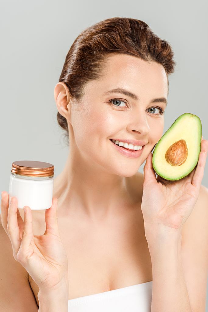 happy woman holding half of avocado and container with cosmetic cream isolated on grey  - Photo, Image