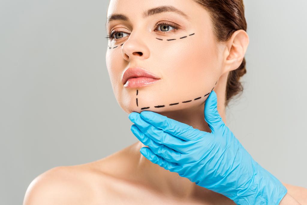 灰色で隔離されたマークされた顔を持つ魅力的な女性の顔に触れる整形外科医のトリミングされたビュー  - 写真・画像