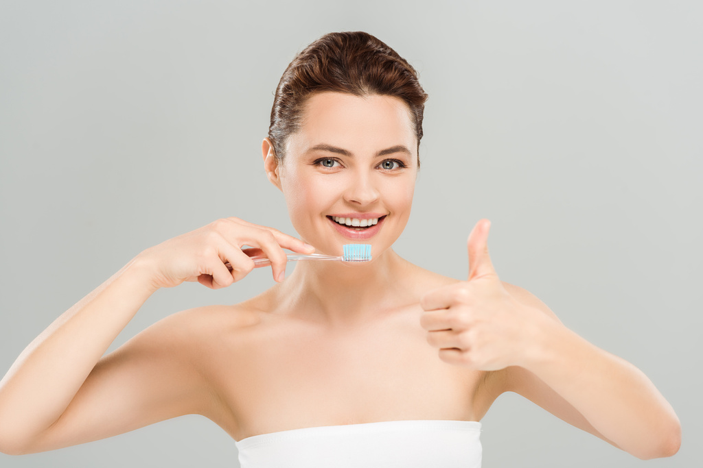 χαρούμενη γυναίκα που δείχνει τον αντίχειρά της κρατώντας οδοντόβουρτσα απομονωμένη σε γκρι  - Φωτογραφία, εικόνα