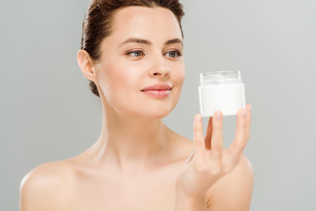 vrolijke naakte vrouw kijkt naar container met cosmetische crème geïsoleerd op grijs  - Foto, afbeelding