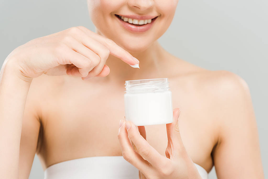 cropped view of smiling woman holding container with face cream isolated on grey  - Photo, Image