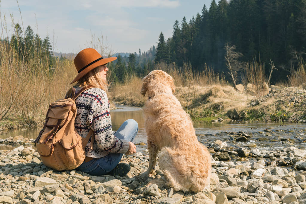 Utazótáska asszony és Arany-Vizsla kutya-ban természet. Stílusos csípő a barna kalap és gyapjú pulóvert a szüreti textil hátizsák közelében hegyek folyó és erdő. Utazás és mehetnék koncepció.  - Fotó, kép