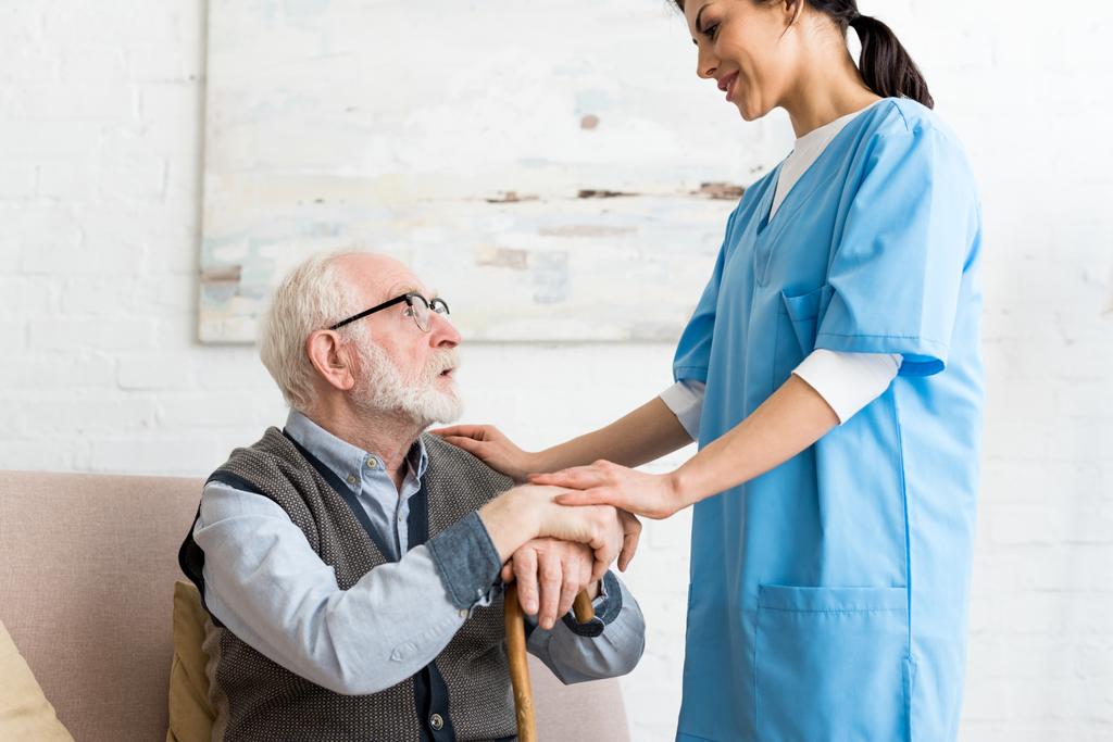 Вид сбоку на пожилого мужчину и медсестру, стоящих в комнате, смотрящих друг на друга
 - Фото, изображение