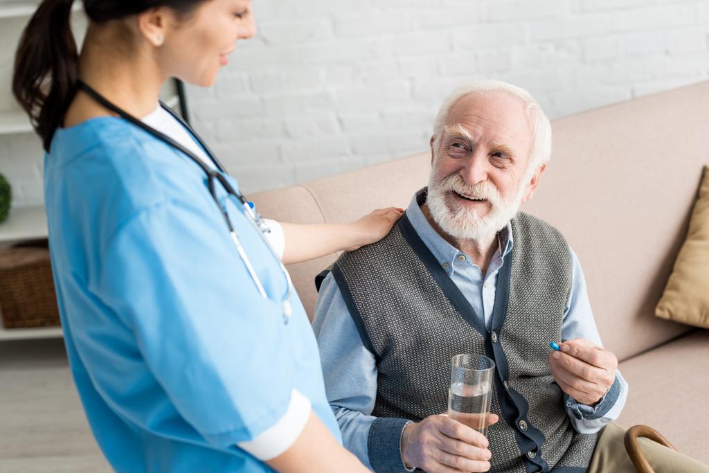Pohled na ošetřovatelku s vysokým úhlem pohledu na šťastného šedovlasého muže s pilulkou - Fotografie, Obrázek