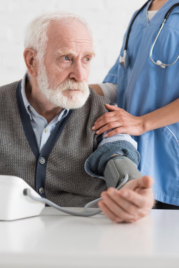 Ausgeschnittene Ansicht einer Krankenschwester, die einem grauhaarigen Mann die Hände aufsetzt und den Blutdruck misst - Foto, Bild