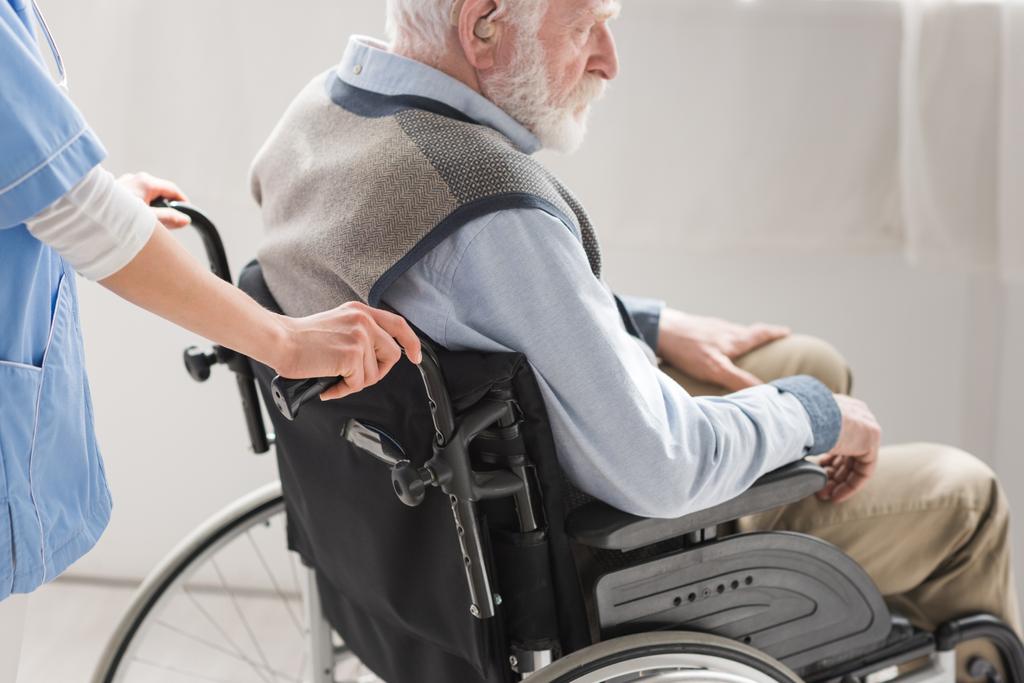 Przycięty widok pielęgniarki stojący za niepełnosprawnym starszym mężczyzną na wózku inwalidzkim  - Zdjęcie, obraz
