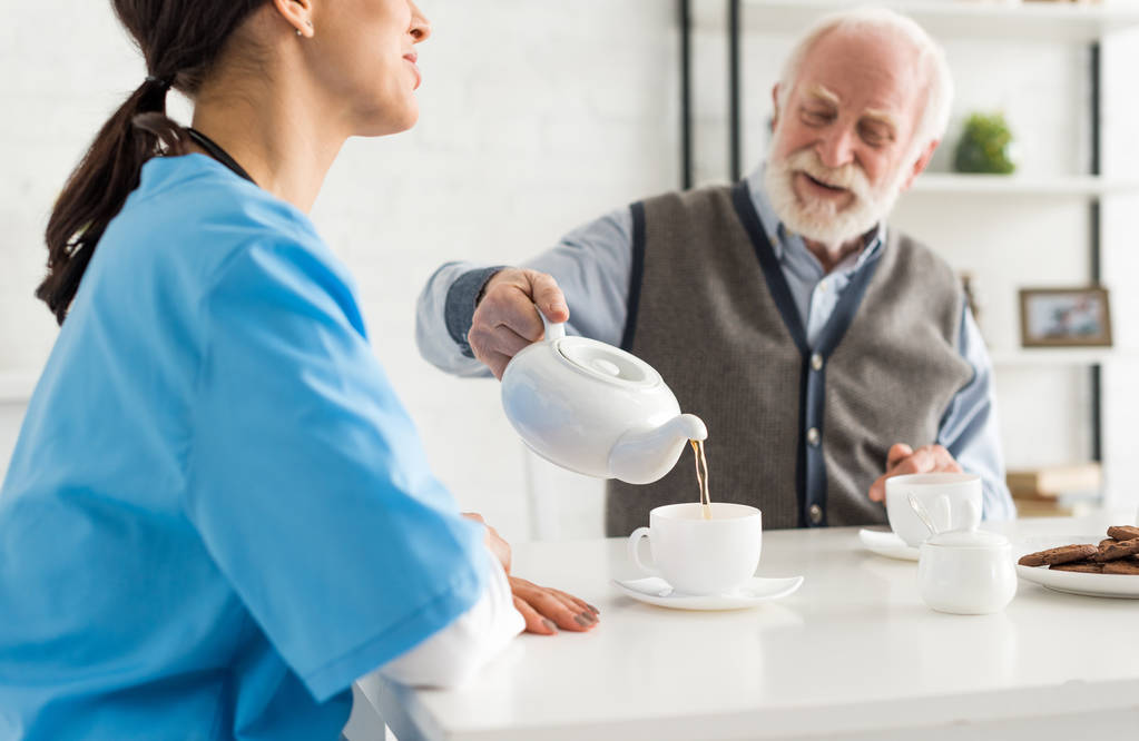 Ευτυχισμένος και χαμογελαστός ανώτερος άνθρωπος, ρίχνει το τσάι σε λευκό Κύπελλο, κάθεται στην κουζίνα στο σπίτι - Φωτογραφία, εικόνα