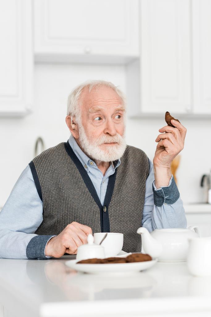 Ciekawy siwy człowiek siedzi na kuchni z ciasteczkiem w ręku, i patrząc z dala - Zdjęcie, obraz