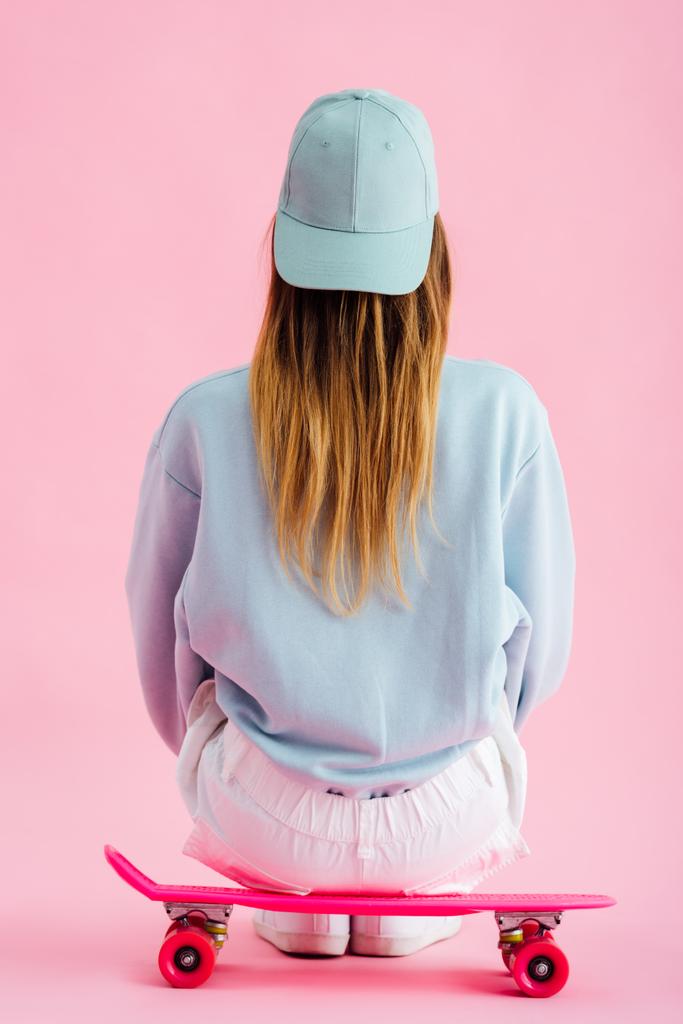 back view of teenage girl in cap sitting on penny board on pink - Photo, Image