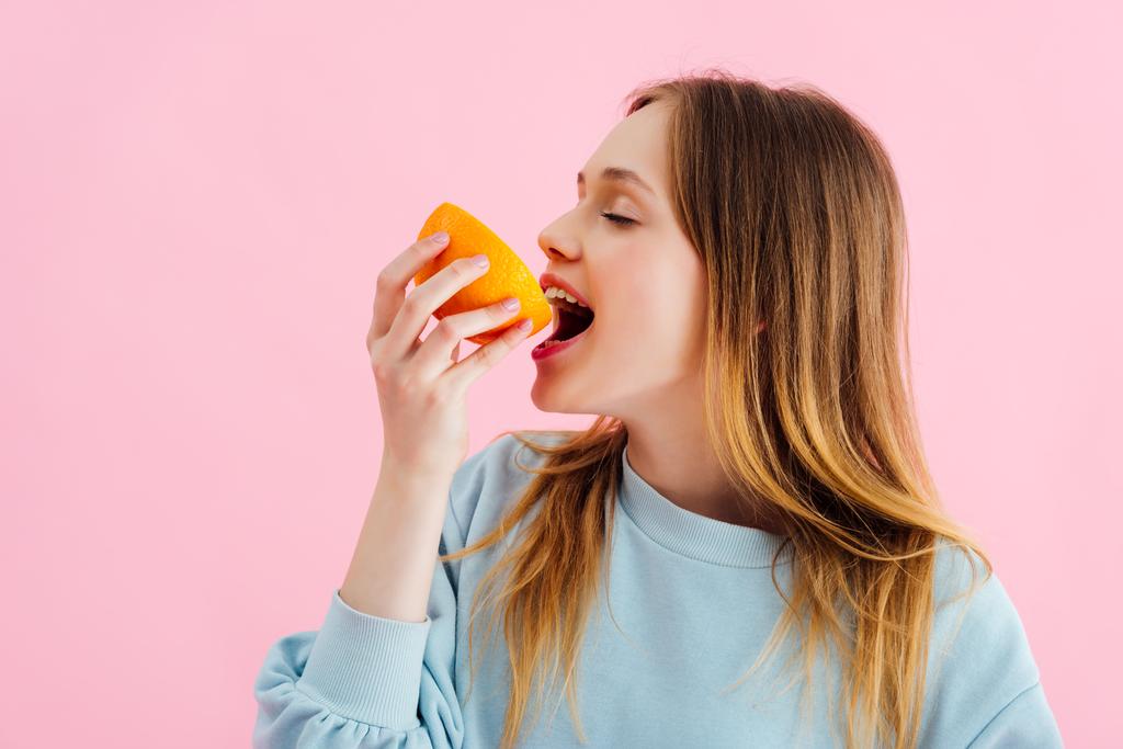 hübsche Teenager-Mädchen mit geschlossenen Augen beißen orange halb isoliert auf rosa - Foto, Bild