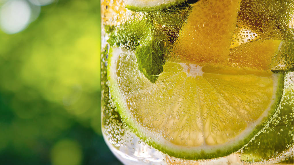 Καλοκαίρι ανθρακούχο κρύο φρέσκο ποτό με φέτες λεμονιού και λάιμ, φύλλο δυόσμου σε ένα γυαλί σε ένα πράσινο φύλλο θαμπό φόντο. Αντιγραφή χώρου. Αλκοολούχα ή μη αλκοολούχα ποτά καλοκαιρινών ποτών. - Φωτογραφία, εικόνα