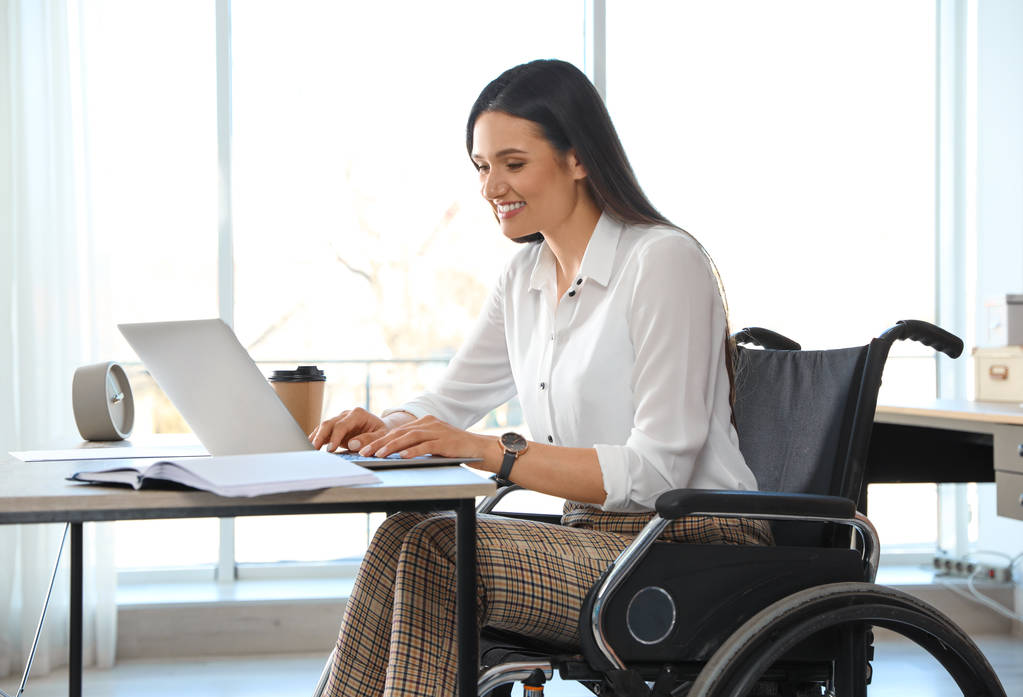 Jovem mulher em cadeira de rodas usando laptop no local de trabalho
 - Foto, Imagem