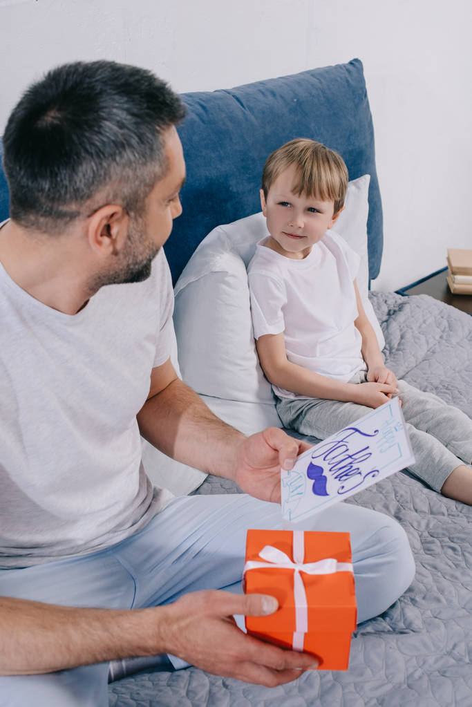 man holding gift box and fathers day greeting card while sitting near adorable son - Photo, Image