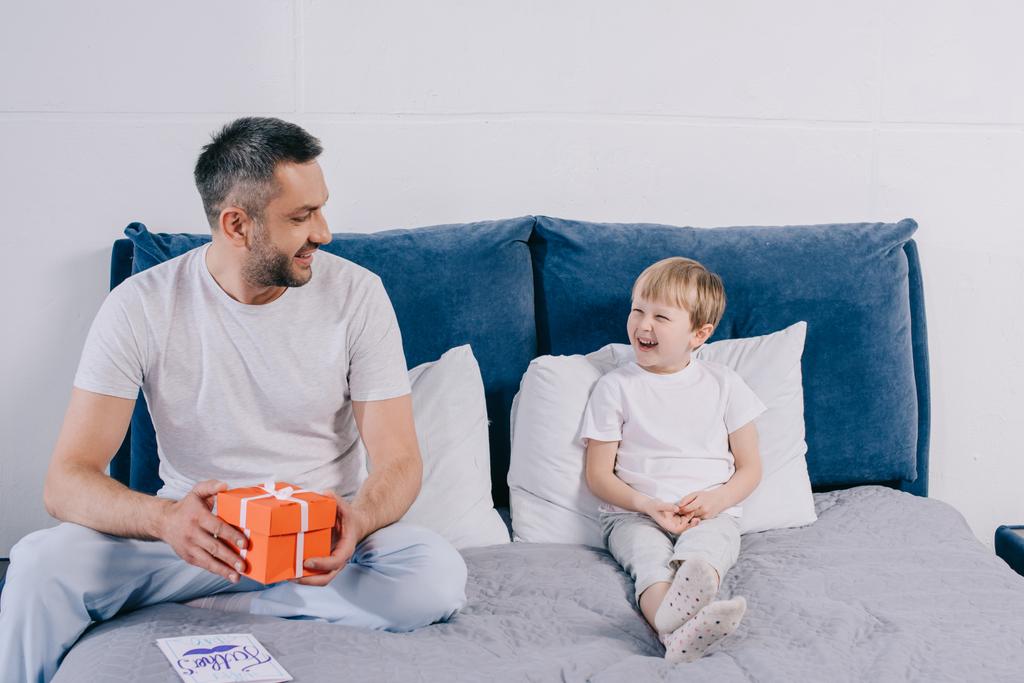 glücklicher Mann, der eine Geschenkbox für den Vatertag in der Hand hält, während er neben seinem fröhlichen Sohn auf dem Bett sitzt - Foto, Bild
