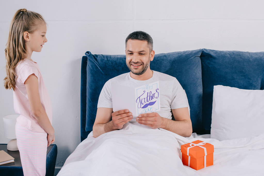 Niedliches Kind steht neben Vater mit Vatertags-Grußkarte im Bett - Foto, Bild