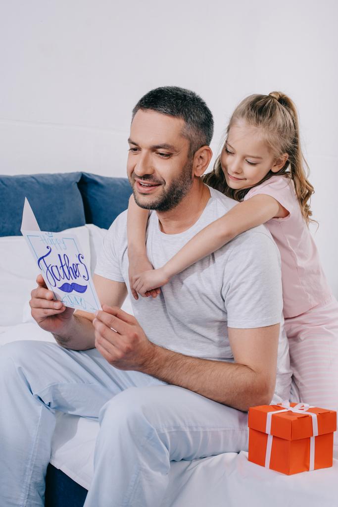 niedliches Kind umarmt glücklichen Vater mit Vatertags-Grußkarte, während es in der Nähe der Geschenkbox sitzt - Foto, Bild