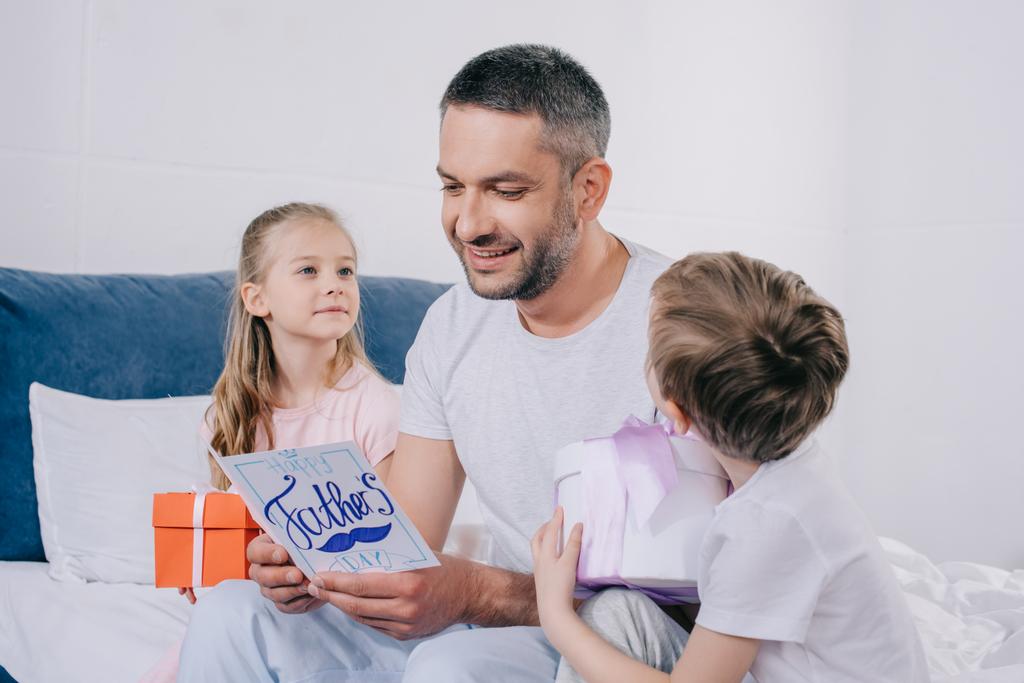 niedliche Tochter und Sohn sitzen mit Geschenkboxen in der Nähe lächelnden Papa hält Vatertags-Grußkarte - Foto, Bild