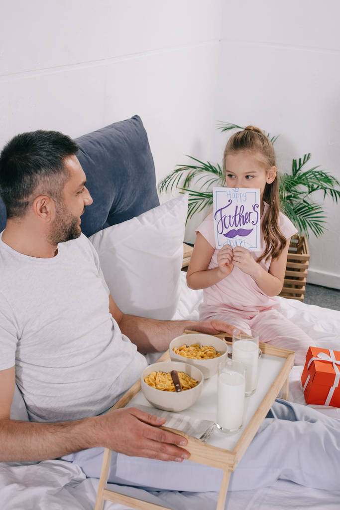 Χαμογελαστός άνθρωπος κρατώντας δίσκο με το πρωινό, ενώ κάθονται στο κρεβάτι κοντά αξιολάτρευτο κόρη καλύπτοντας το πρόσωπο με ευχετήρια κάρτα ημέρα του πατέρα - Φωτογραφία, εικόνα
