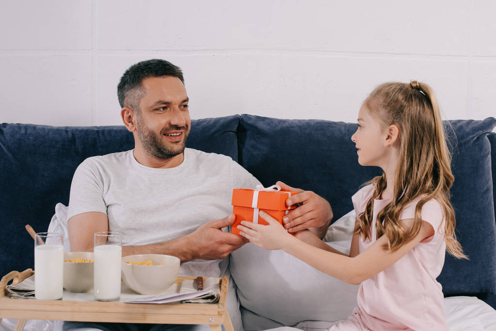 adorable daughter presenting fathers day gift box to smiling dad having breakfast in bed - Photo, Image