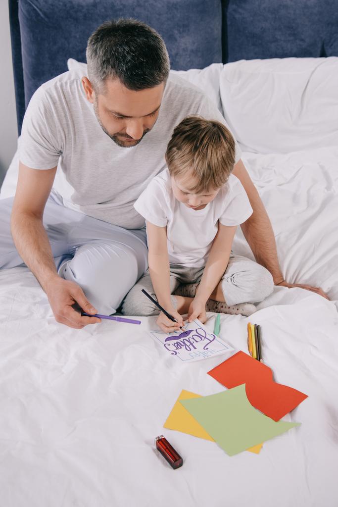 милий син малює батьків день вітальна листівка сидячи на постільній білизні біля тата
 - Фото, зображення