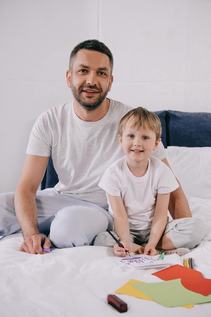 愛らしい息子の近くに寝具の上に座って幸せな男は父親の日のグリーティングカードを作ります - 写真・画像