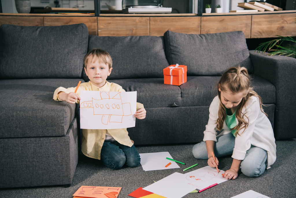 cute boy showing drawing while sitting on floor near sofa together with sister - Photo, Image