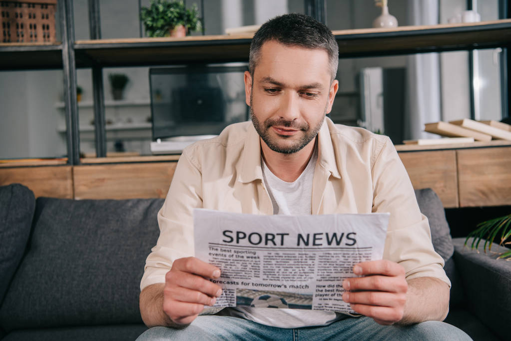 όμορφος άνθρωπος ανάγνωση αθλητικών ειδήσεων εφημερίδα, ενώ κάθεται στον καναπέ στο σπίτι - Φωτογραφία, εικόνα