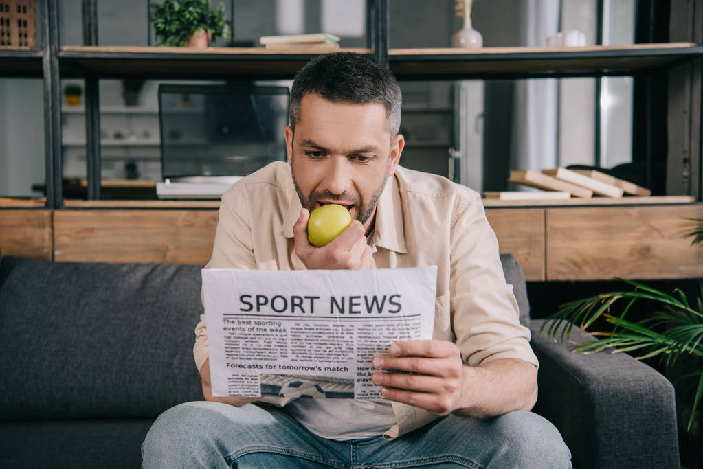 schöner Mann isst frischen Apfel, während er zu Hause Sportnachrichten liest - Foto, Bild