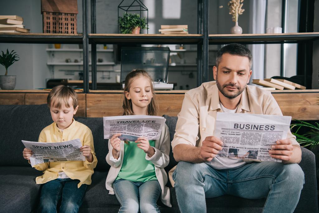 father with adorable kids reading newspapers while sitting on sofa at home - Photo, Image