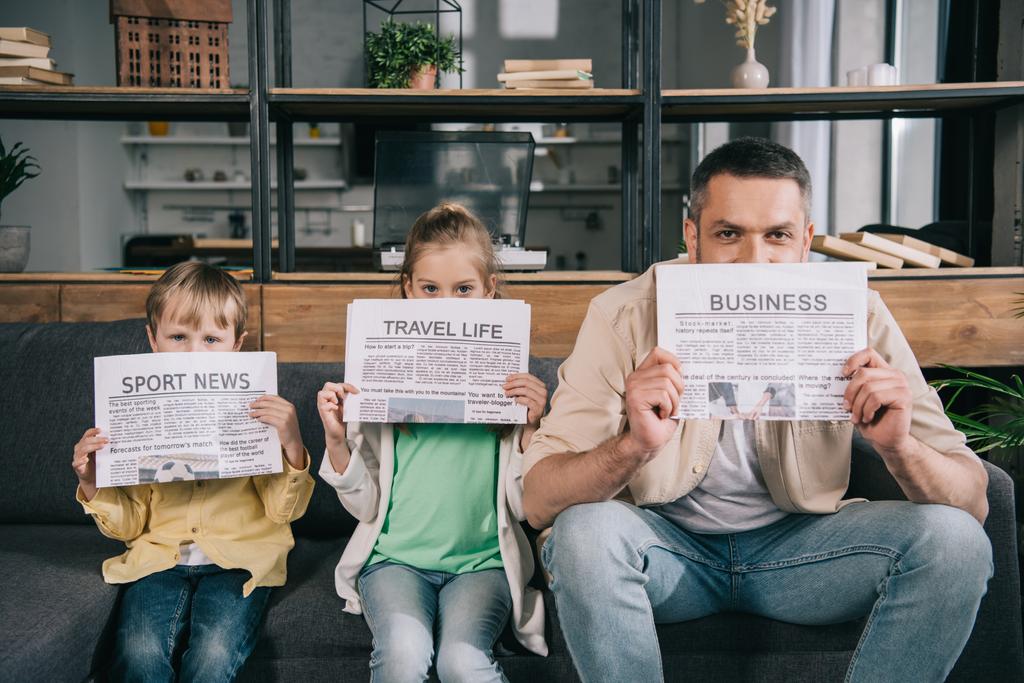 cheerful father and kids holding newspapers while sitting on sofa at home - Photo, Image