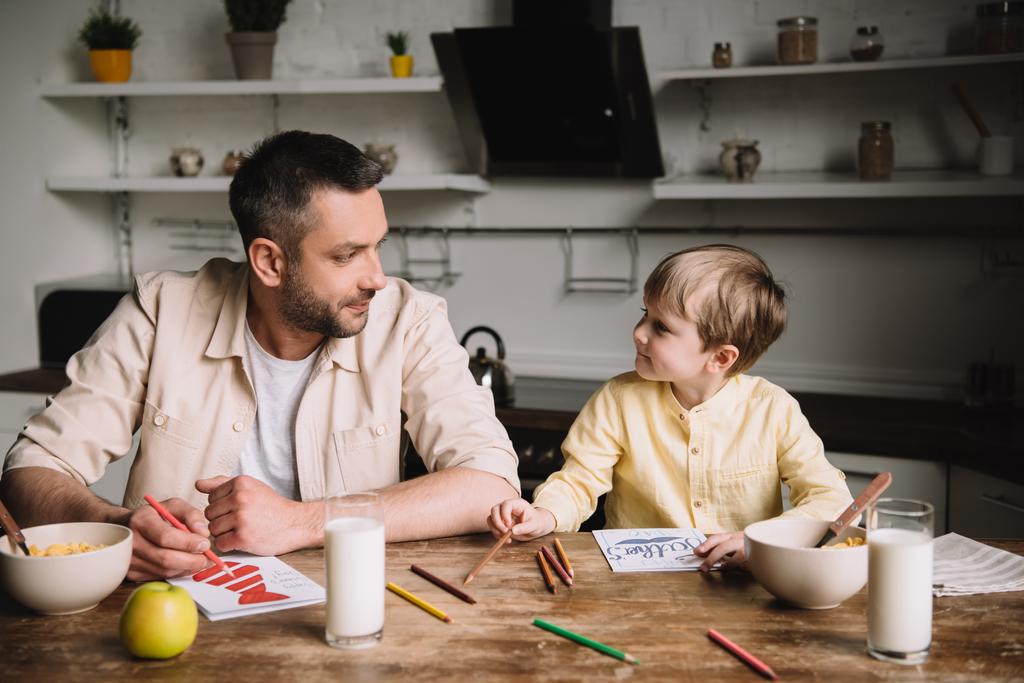 father and son drawing while sitting at kitchen table with served breakfast - Photo, Image