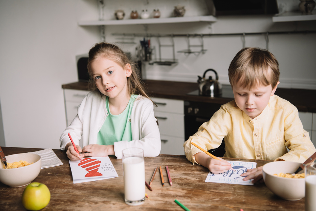 adorable hermano y hermana sentado en la mesa de la cocina con el desayuno servido y dibujar tarjetas de felicitación día padres
 - Foto, imagen
