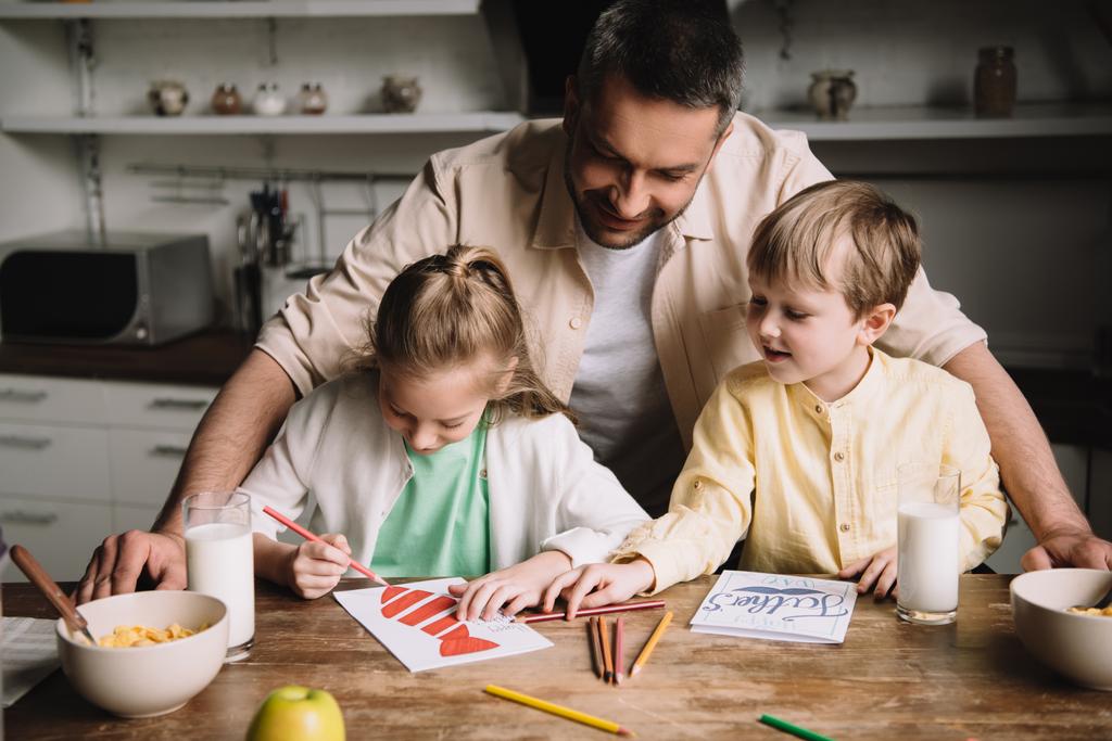 feliz papá de pie cerca adorable niños dibujo padres día tarjetas de felicitación mientras está sentado en la mesa de la cocina con el desayuno servido
 - Foto, imagen