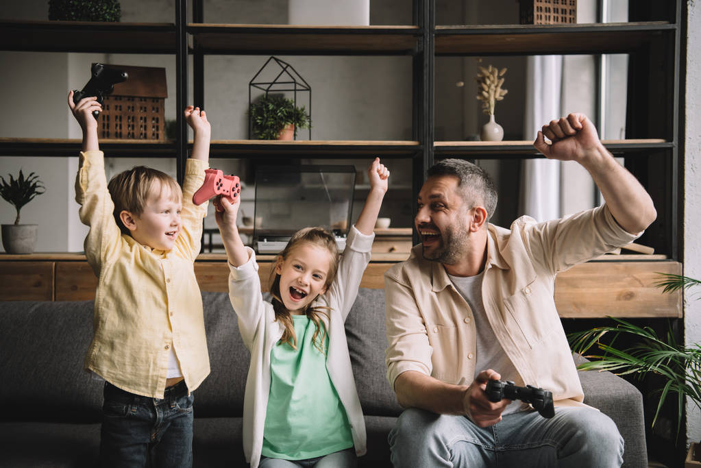 KYIV, UCRANIA - 10 DE MAYO DE 2019: Feliz familia celebrando el triunfo mientras juega videojuegos con joysticks en casa
.  - Foto, imagen