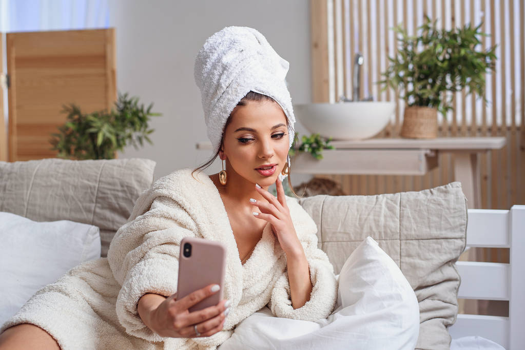 Sensual jovem mulher em roupão de banho e com toalha na cabeça fazendo selfie usando smartphone na cama em casa aconchegante
. - Foto, Imagem