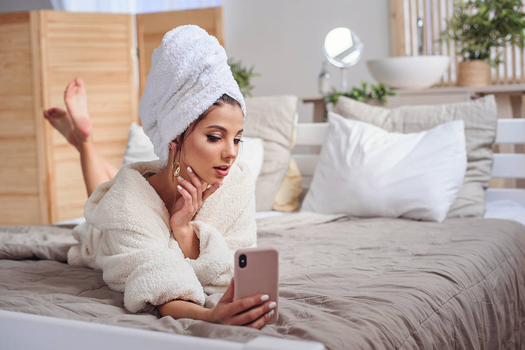 Красива дівчина в халаті і з рушником на голові робить селфі, використовуючи смартфон на ліжку в затишному готельному номері
. - Фото, зображення