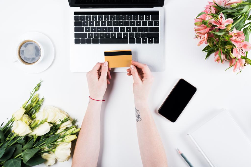 vista cortada de mulher segurando cartão de crédito perto de laptop, smartphone com tela em branco, xícara de café e buquês no branco
 - Foto, Imagem