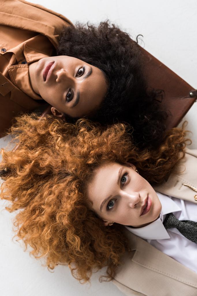 Top uitzicht van Redhead meisje liggend op koffer met Afro-Amerikaanse krullend vrouw geïsoleerd op wit e  - Foto, afbeelding
