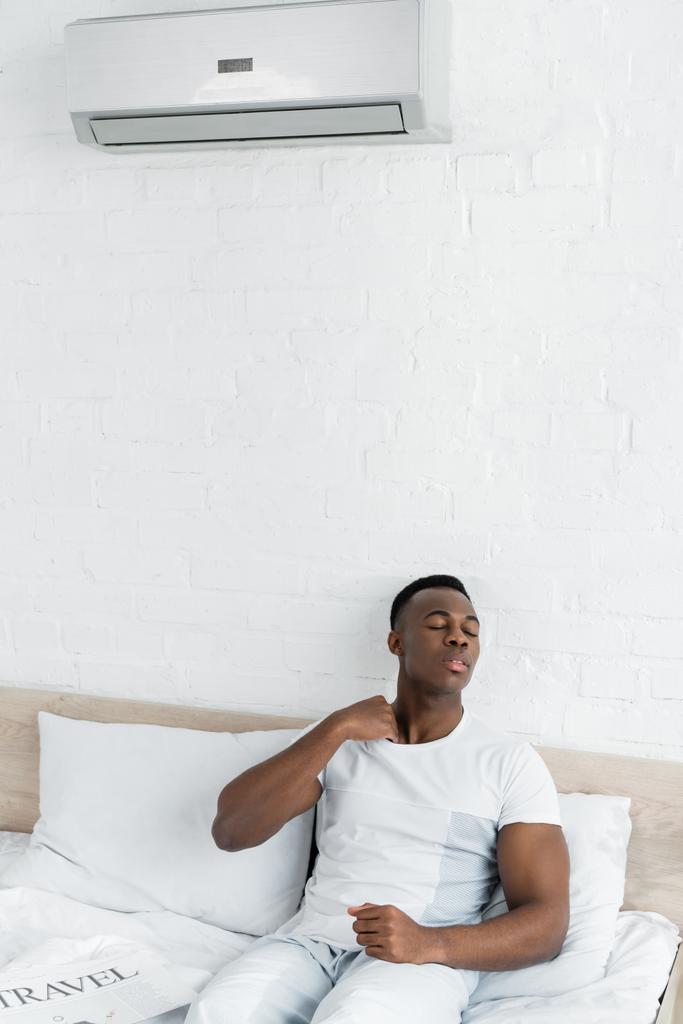 ιδρωμένος Αφρικανός Αμερικανός άνθρωπος κάθεται στο κρεβάτι στο δωμάτιο με κλιματισμό - Φωτογραφία, εικόνα